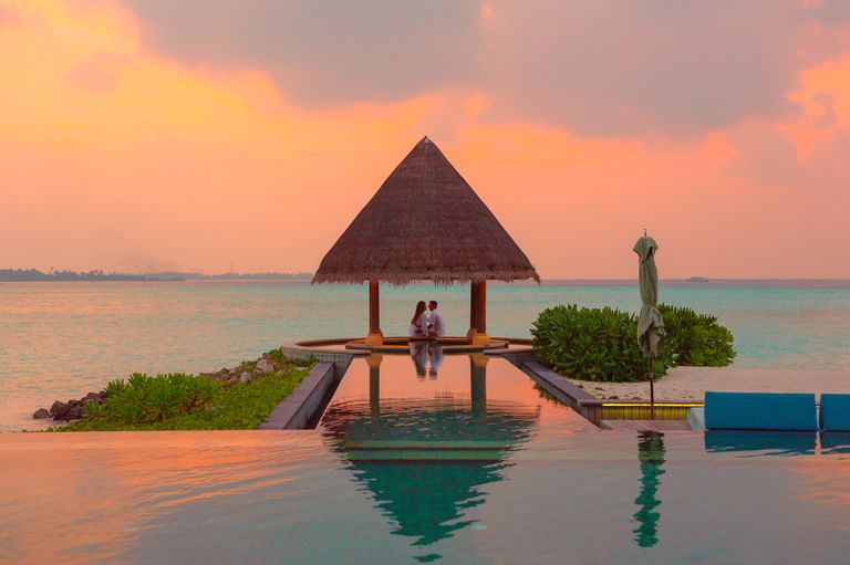 A couple enjoying a romantic moment by the sea and pool under a beach cabana during sunset on a luxury travel itinerary.